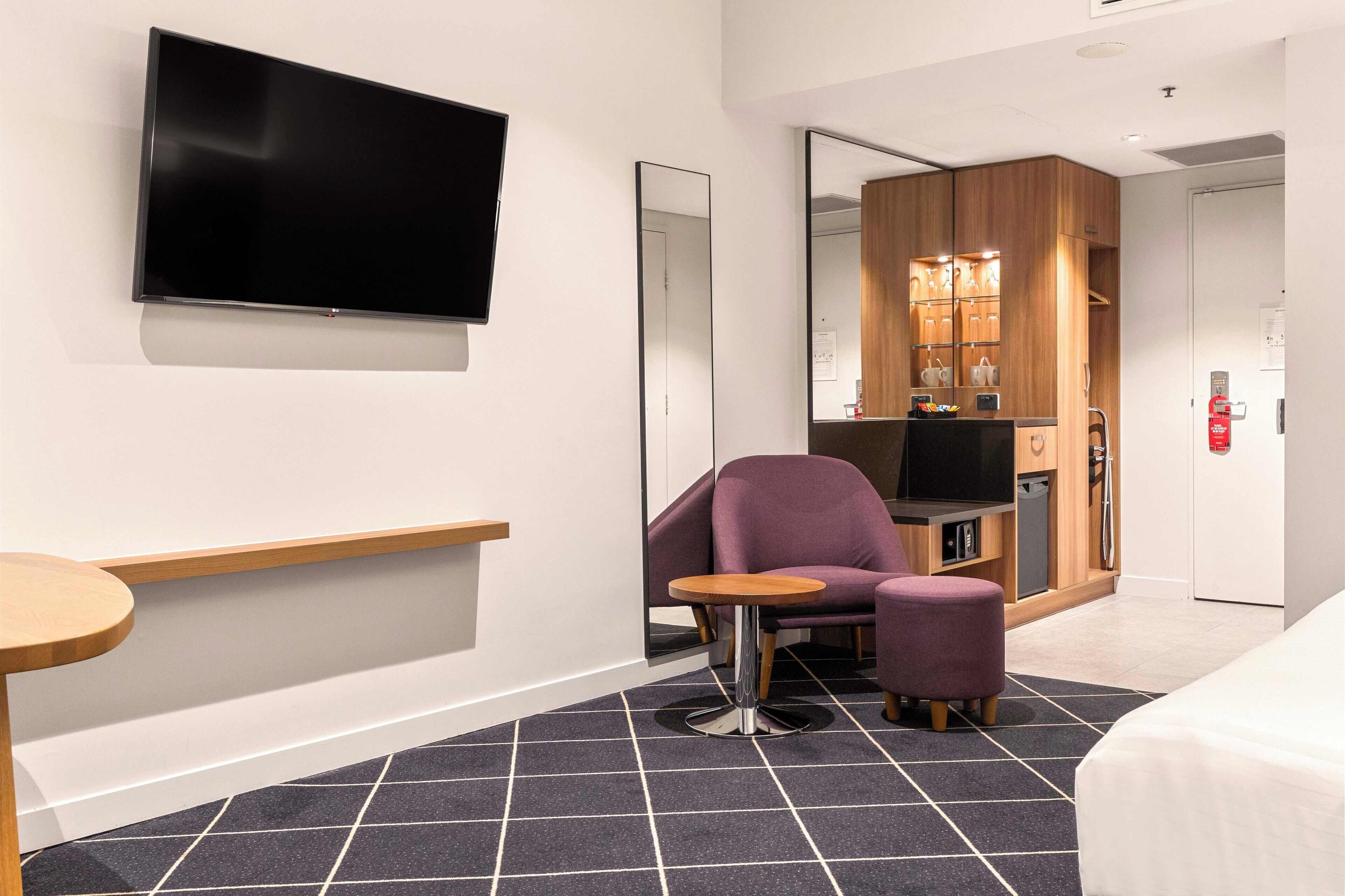 A modern hotel room with a flat-screen TV, a large mirror, a wooden cabinet, a purple armchair, a small round table, and a bed.