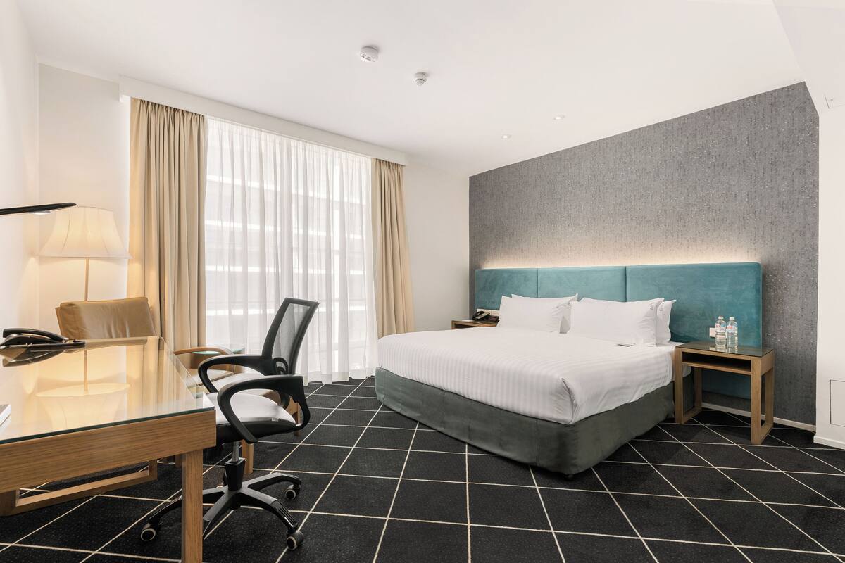 Ein Hotelzimmer mit Bett, Schreibtisch mit Stuhl, Nachttisch und einem Fenster mit Vorhängen.