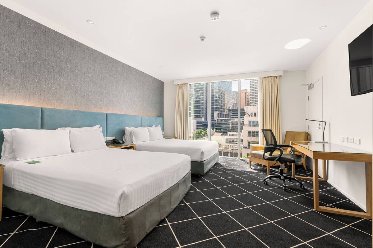 Ein Hotelzimmer mit zwei Betten, einem Schreibtisch und einem großen Fenster mit Blick auf die Stadt.