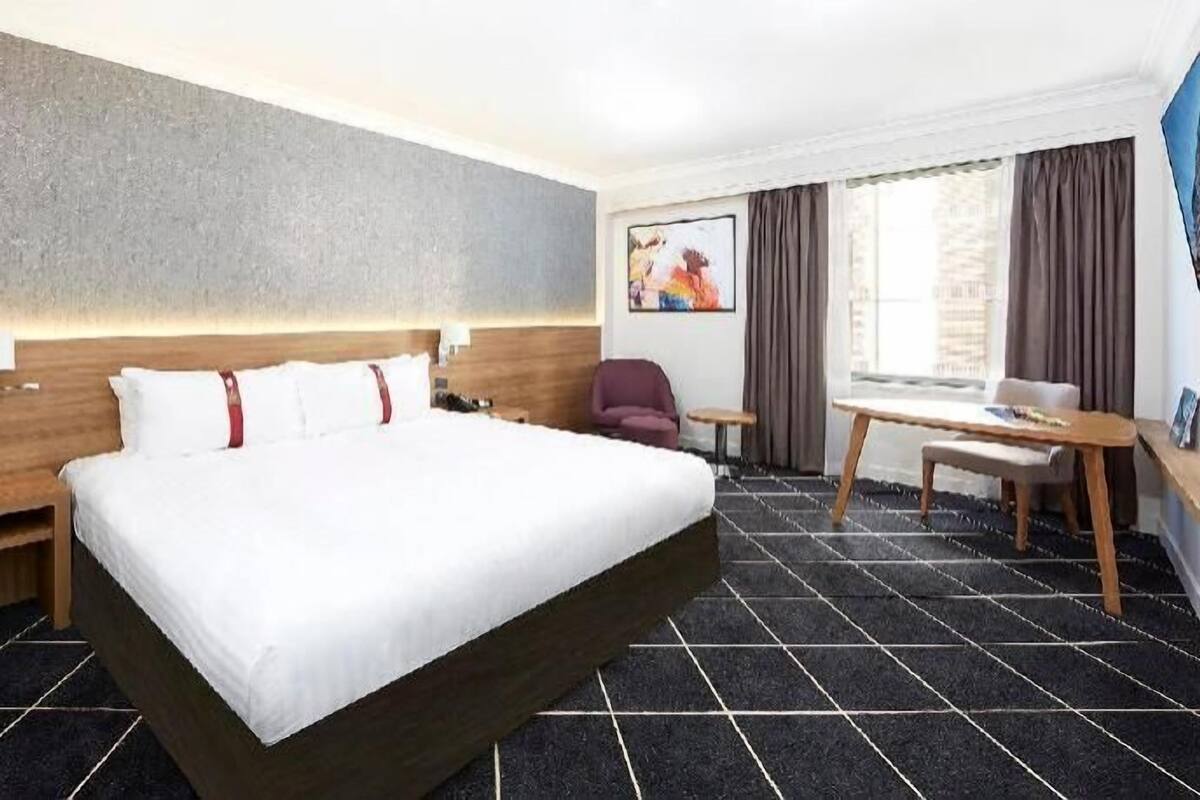 Ein Hotelzimmer mit einem großen Bett, einem Schreibtisch, einem Sessel und einem Fenster mit Vorhängen.