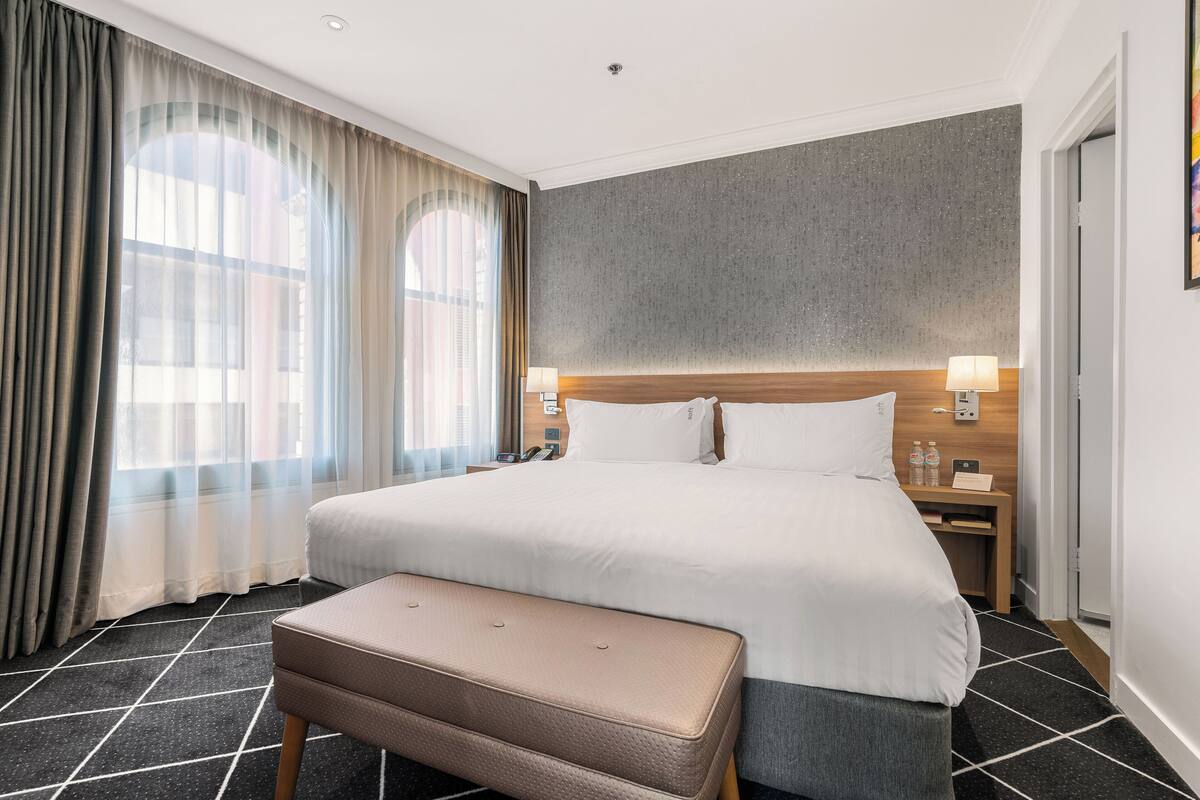 Ein Hotelzimmer mit einem großen Bett, einem Nachttisch mit Lampe, einer Bank und Blick durch Vorhänge auf die Außenwelt.