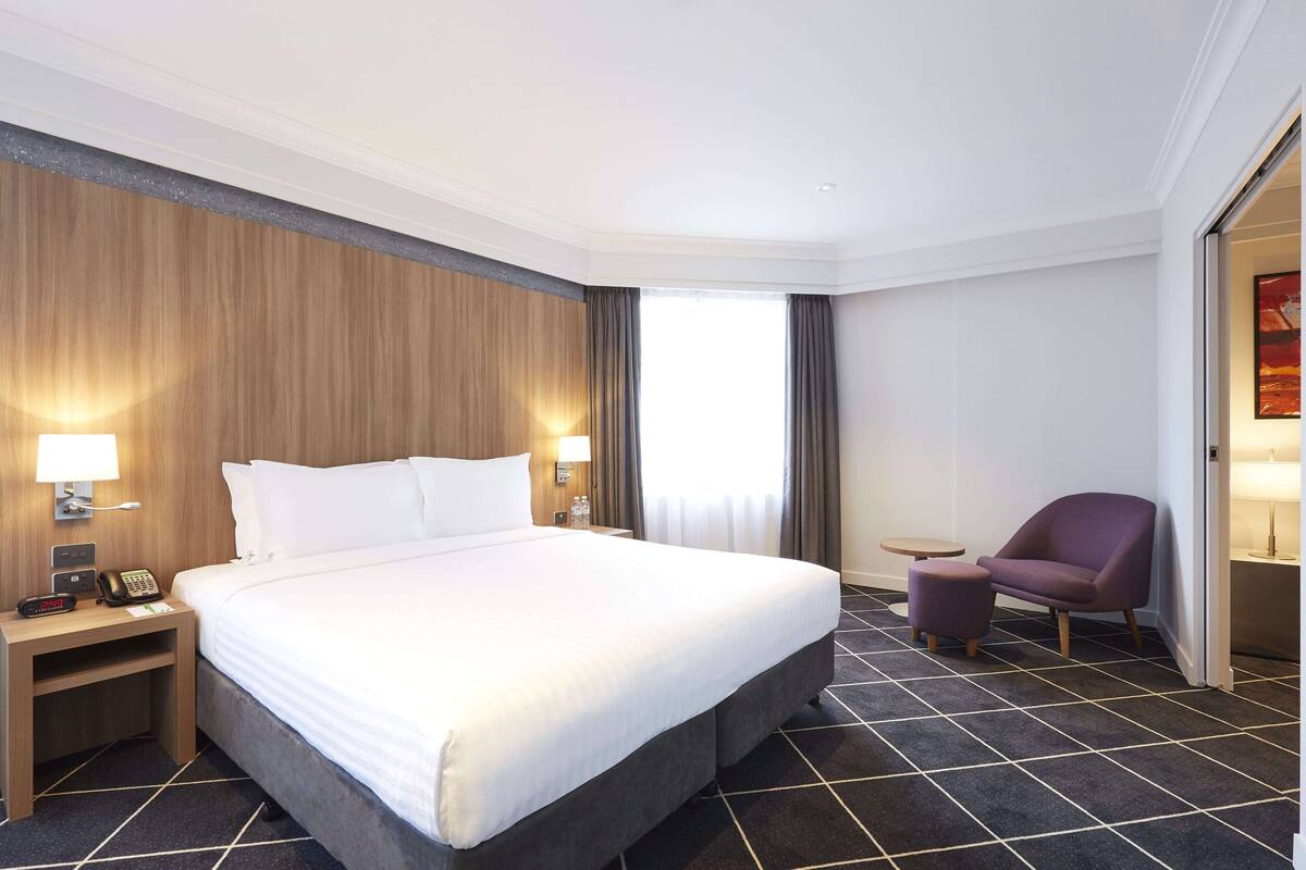 Ein Hotelzimmer mit einem großen Bett, Nachttischlampen, einem kleinen Tisch, einem Sessel und Blick durch ein Fenster nach draußen.