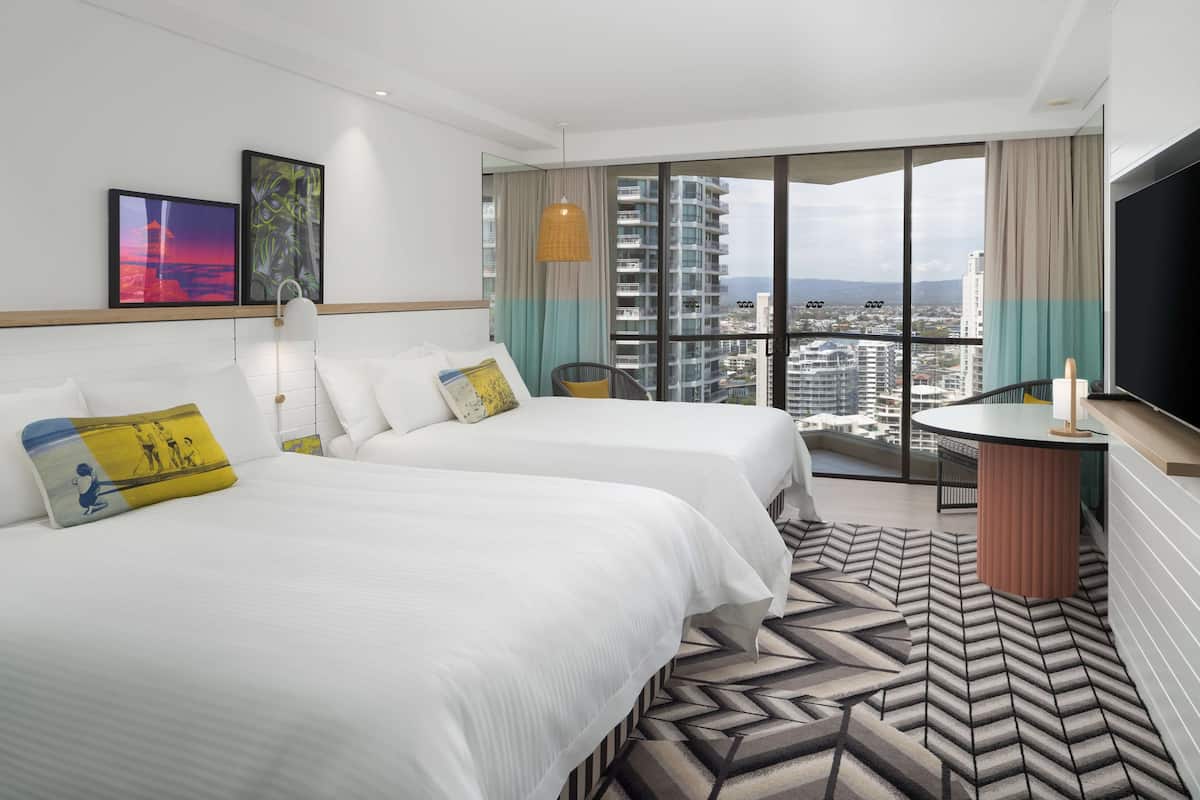 A hotel room with two beds, a large window with a city view, a flat-screen TV, and a patterned rug.