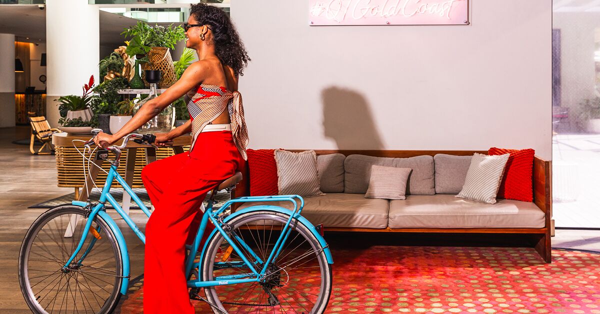 A woman riding a blue bicycle in a modern indoor setting with a sofa and decorative plants in the background.
