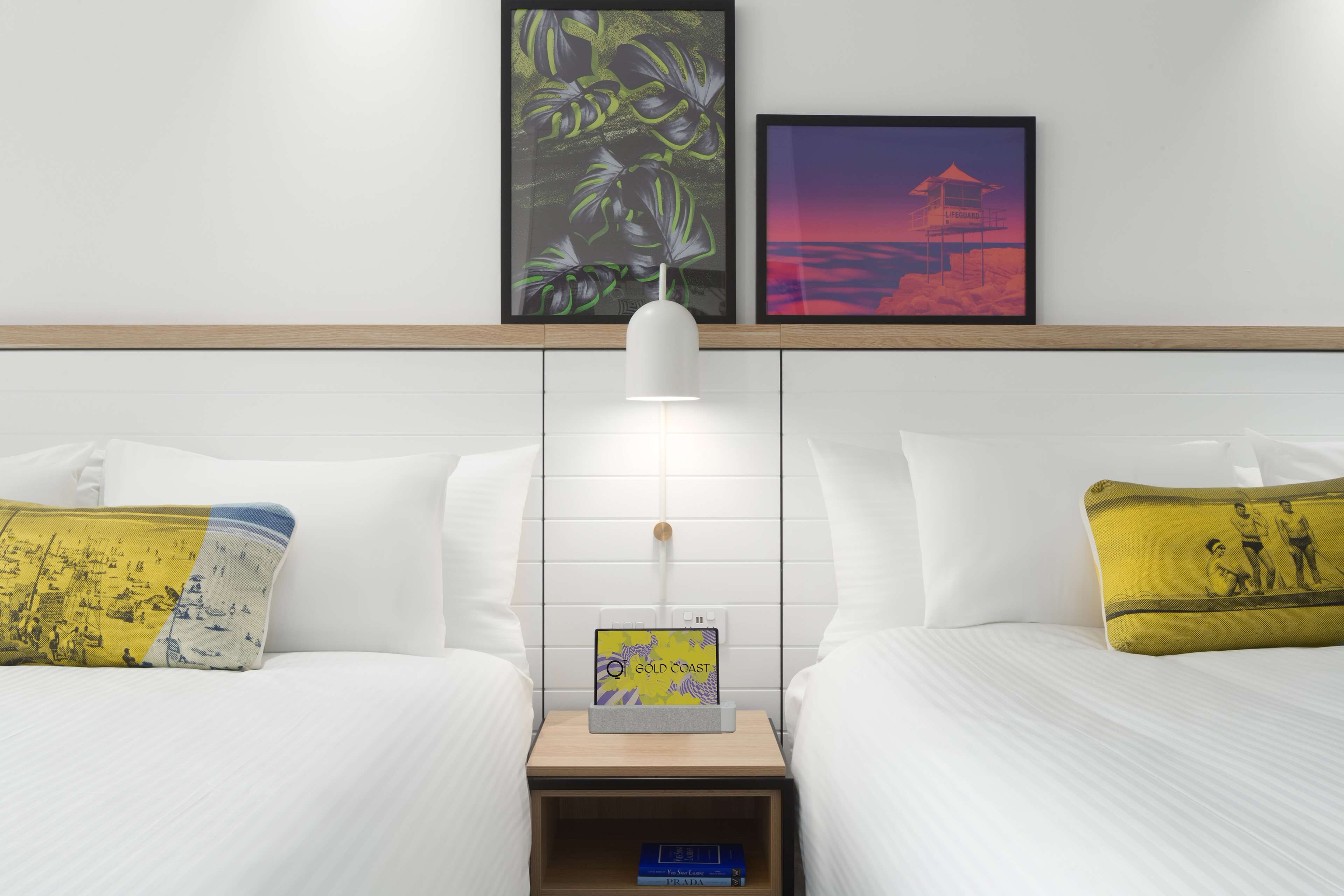 A hotel room with two beds, a nightstand with a lamp, a small table, and two framed pictures on the wall.