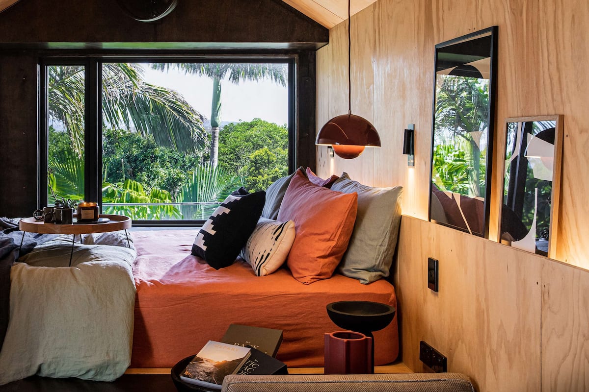 A cozy bedroom with a large window, a bed with multiple pillows, a side table with a tray, and a wooden wall.