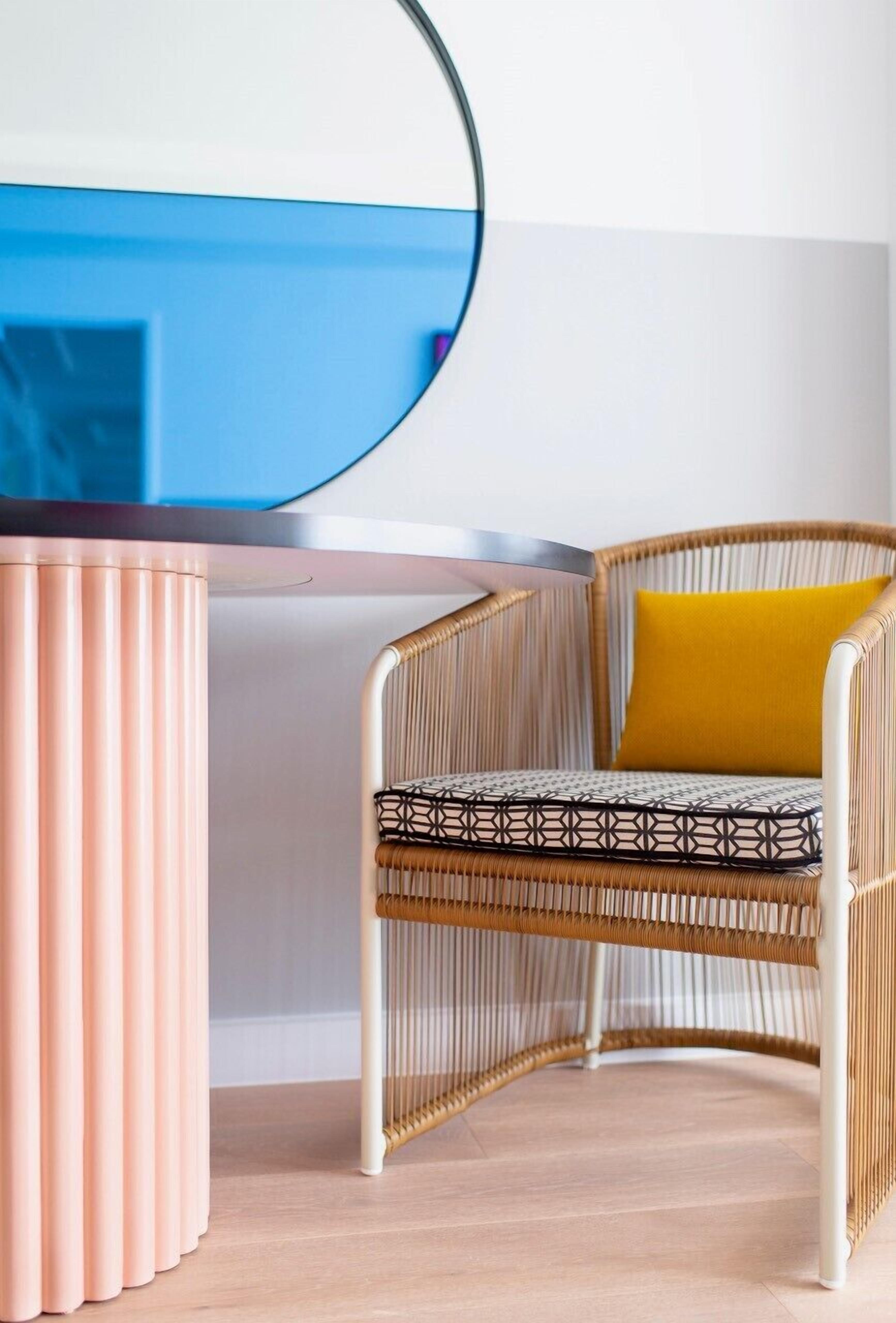 A wicker chair with a geometric patterned cushion and a mustard yellow pillow, positioned next to a pink cylindrical object and a round mirror on a light wooden floor.