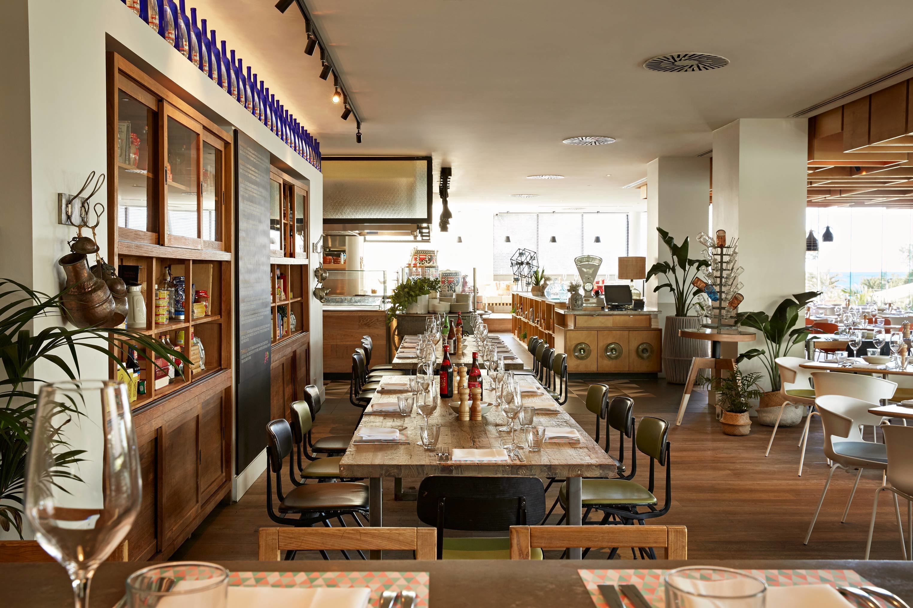 A spacious dining area with a long wooden table set for a meal, surrounded by chairs. There are shelves with various items on the left side, and a counter with a display of bottles and glasses.
