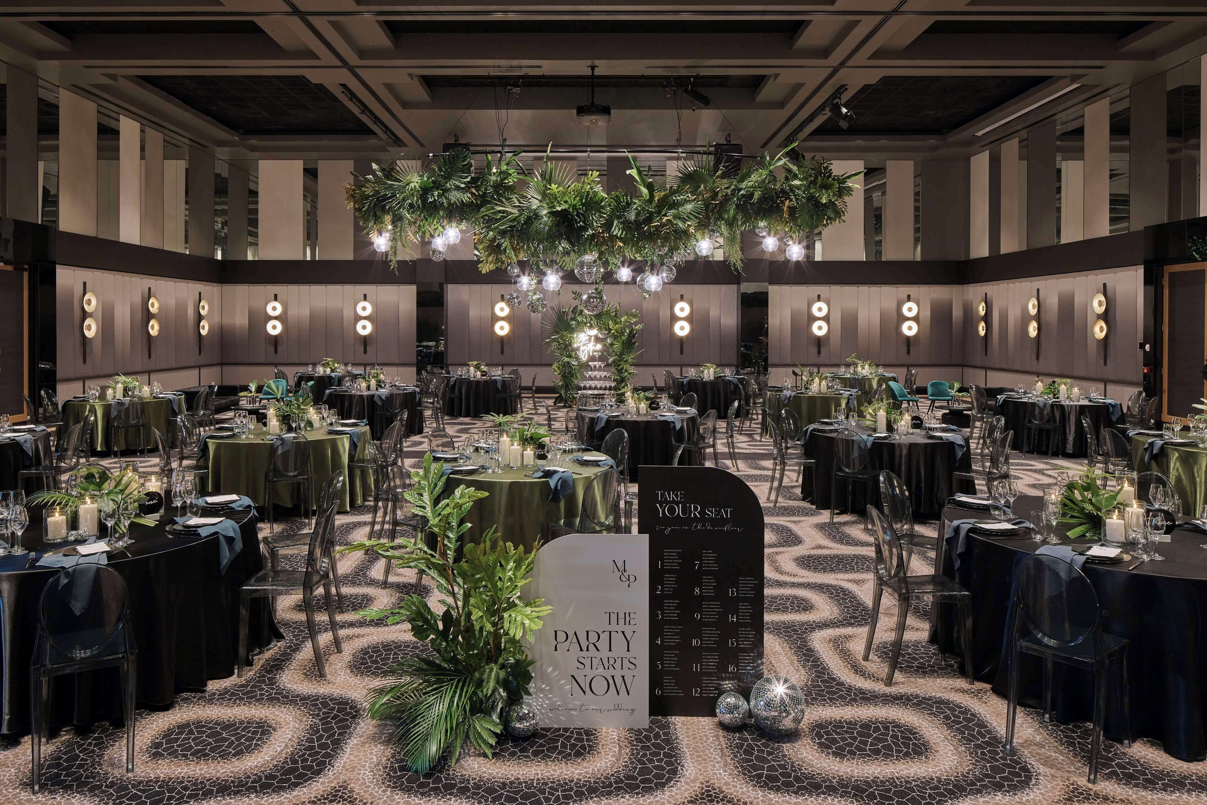 A banquet hall with round tables, clear chairs, and a central floral arrangement. A sign indicates seating options.