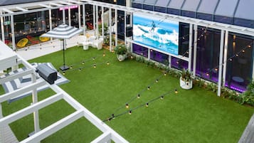 Un jardín en la azotea con césped artificial, mobiliario exterior y una pantalla grande que muestra una escena de surf.