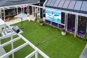 Un jardín en la azotea con césped artificial, mobiliario exterior y una pantalla grande que muestra una escena de surf.