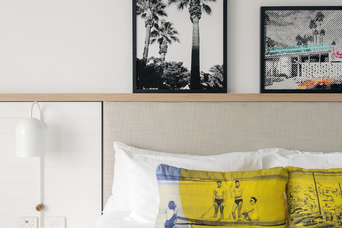 A neatly arranged hotel room with a bed, a framed picture of palm trees, a lamp, and a power outlet.