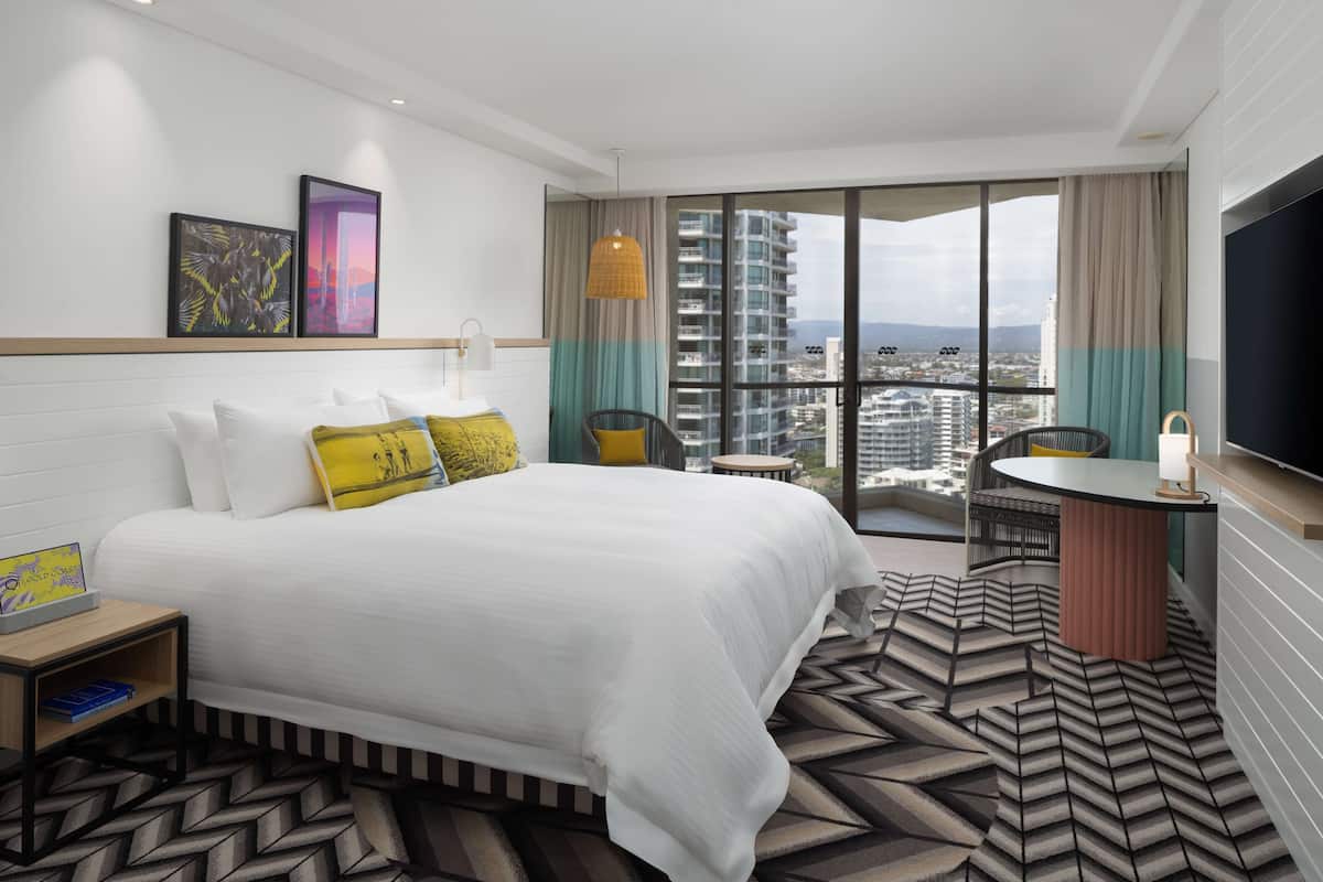 A hotel room with a large bed, a small table, a TV, and a balcony with a city view.