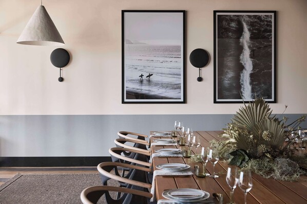 Un espace repas avec une longue table en bois, dressée pour six personnes, flanquée de deux haut-parleurs noirs fixés au mur et agrémentée d’œuvres d’art encadrées sur le thème de l’océan.