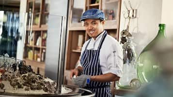 A person in a kitchen setting, wearing a chef's hat and apron, handling food on a counter with various kitchen items and a large green bottle in the background.