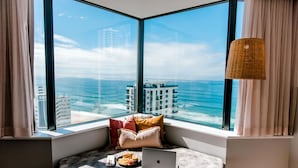 A corner room with a large window offering a view of the ocean and buildings. There is a laptop, a bowl of fruit, and a small table with cushions.