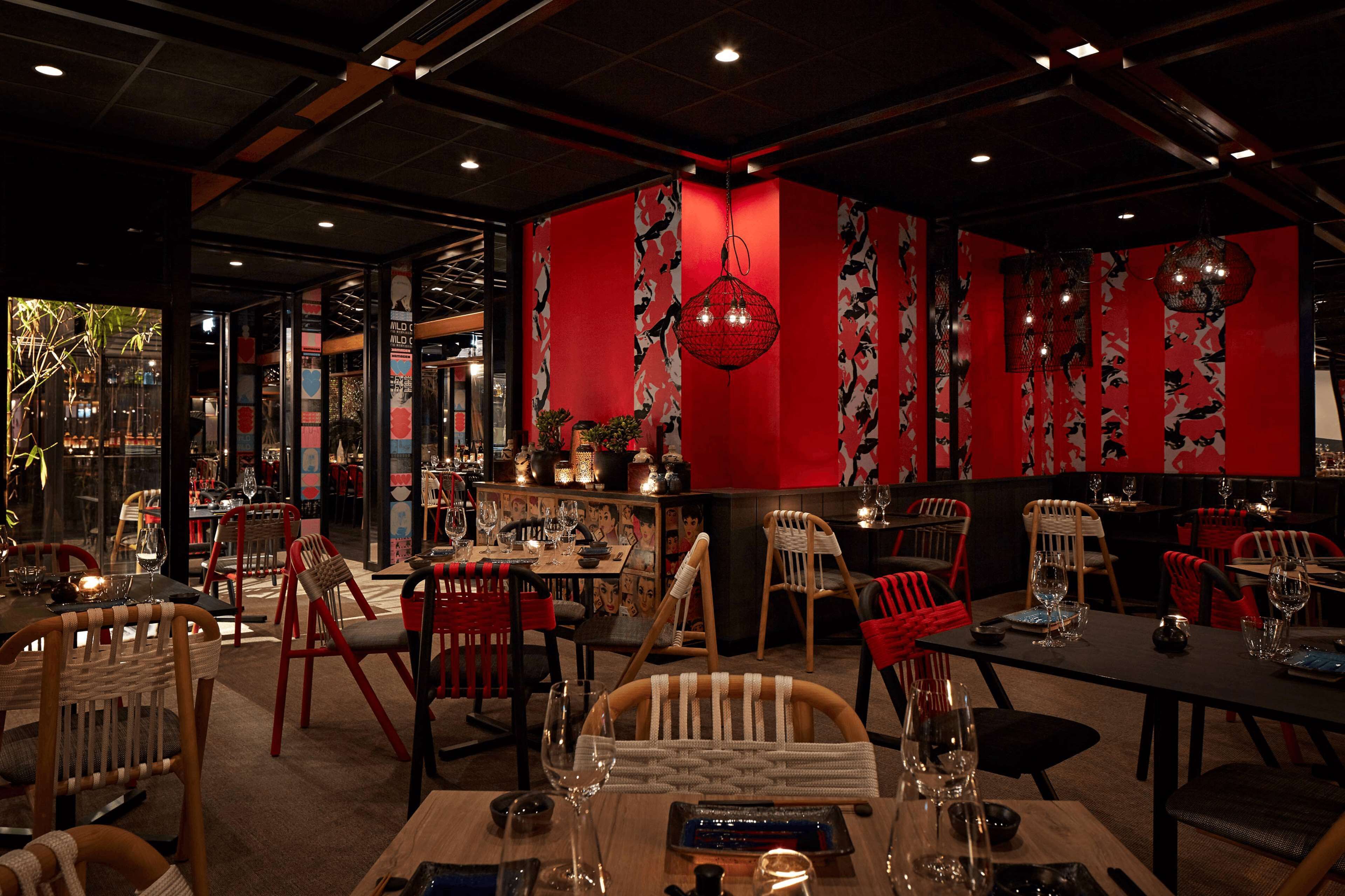 Interior de restaurante con paredes rojas, sillas de madera y faroles colgantes. Las mesas están dispuestas con vasos y cubiertos.