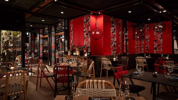 Interior de restaurante con paredes rojas, sillas de madera y faroles colgantes. Las mesas están dispuestas con vasos y cubiertos.