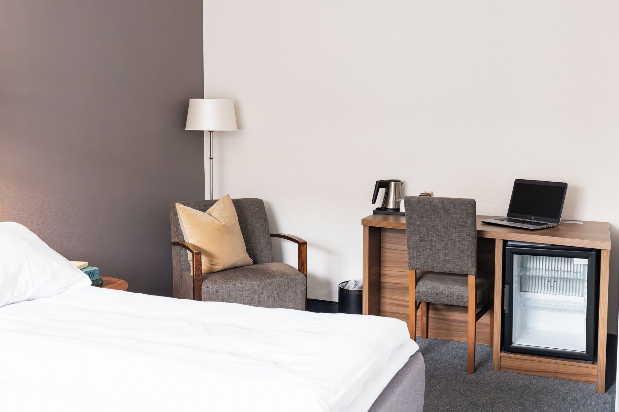 A hotel room with a bed, a desk with a laptop, a chair, a lamp, and a mini-fridge.