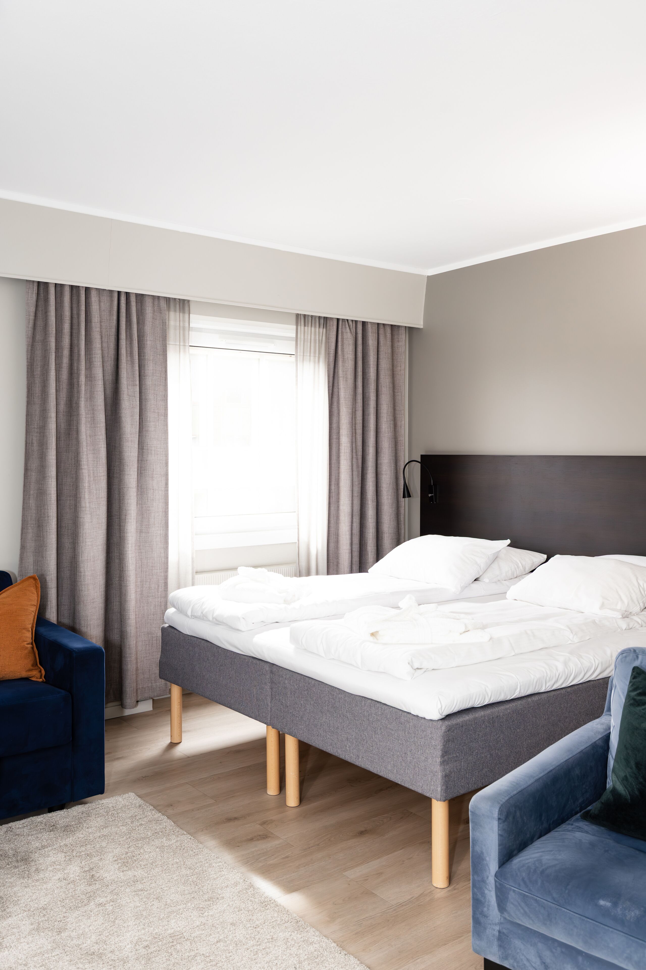 Habitación de hotel moderna con una cama grande, cortinas grises, un sillón azul y piso de madera.