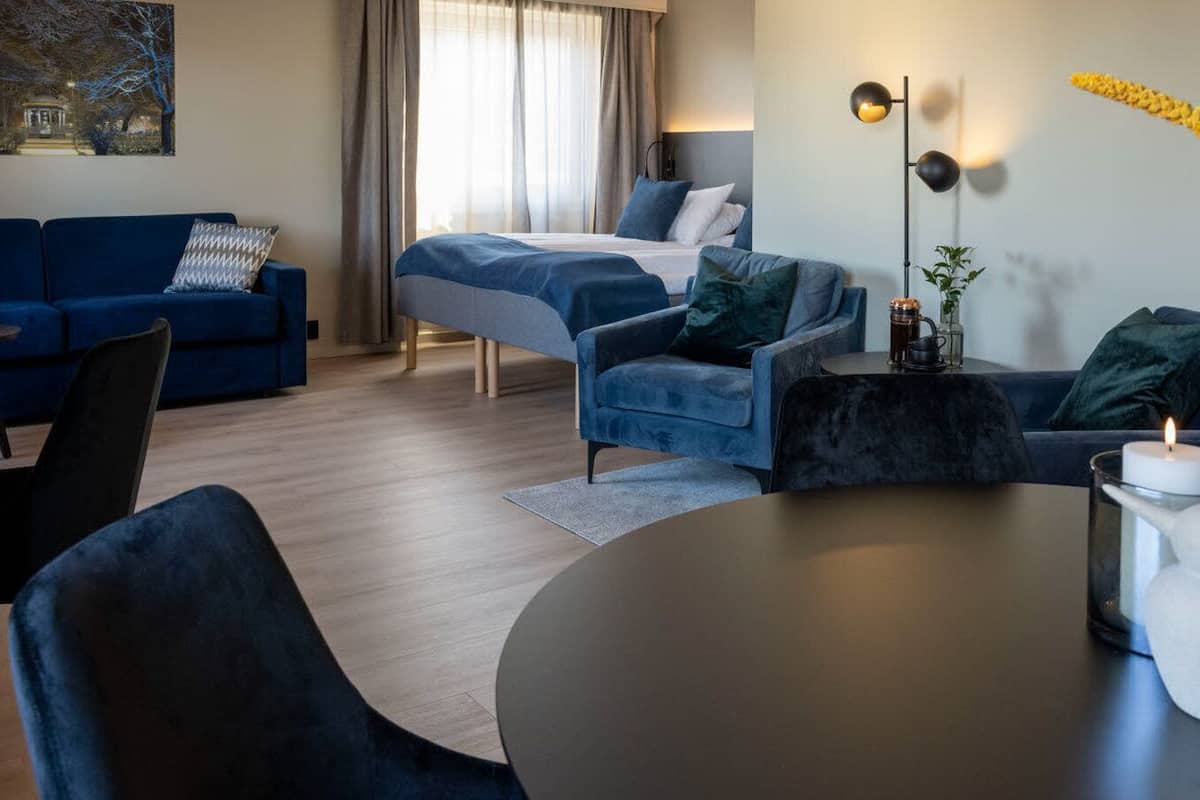 Moderni hotellihuone, jossa on sänky, kaksi sohvaa, ruokapöytä ja sohvapöytä. Seinällä on taulu ja pöydällä maljakko, jossa on kukkia.