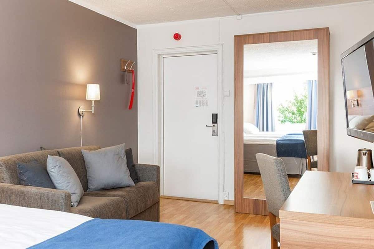 Hotellihuoneessa on sänky, sohva, pöytä ja tuoli. Huoneesta on ovi toiseen huoneeseen.