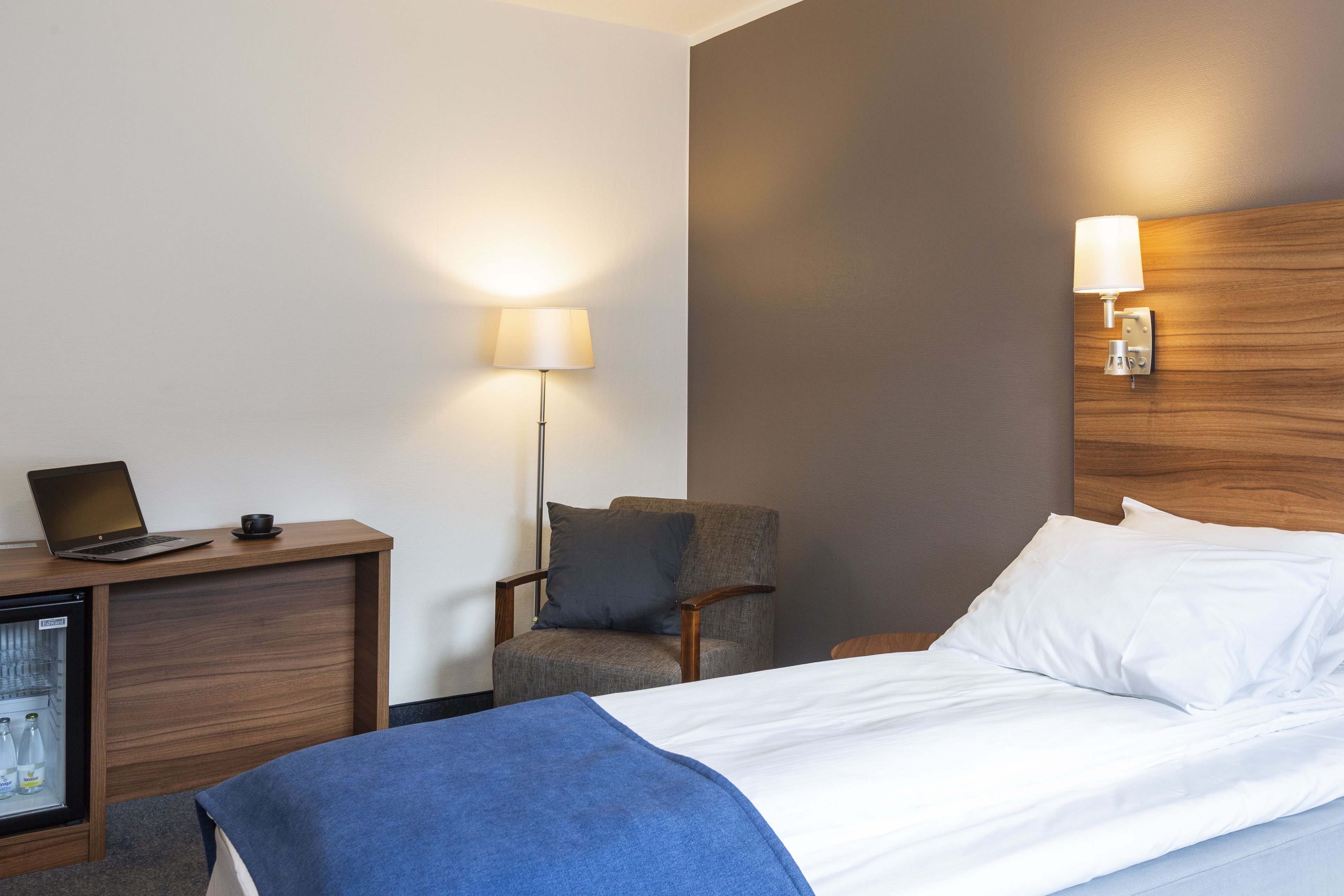 A hotel room with a bed, a desk with a laptop, a chair, a wall-mounted lamp, and a mini-fridge.