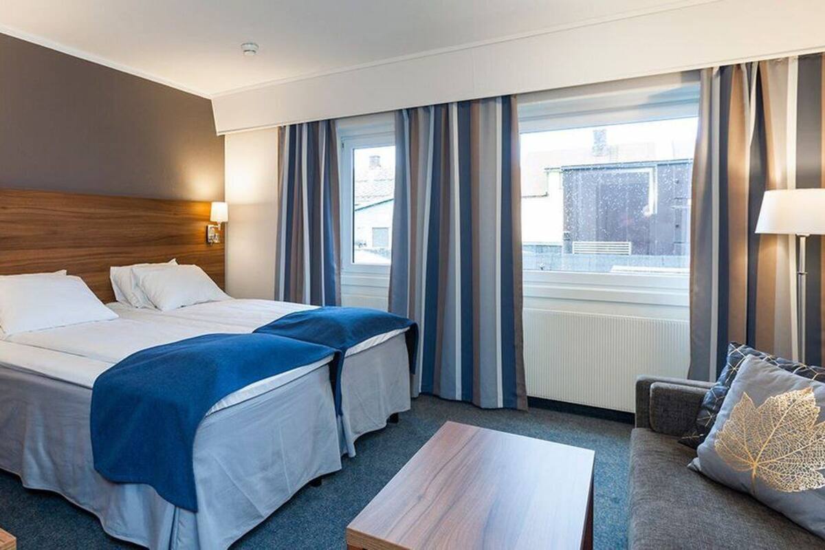 Hotellihuone, jossa on kaksi sänkyä, sohva, pöytä ja ikkuna, jossa on verhot.