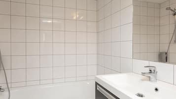 Un baño con baldosas blancas, una bañera, un lavamanos con grifo y un cabezal de ducha.