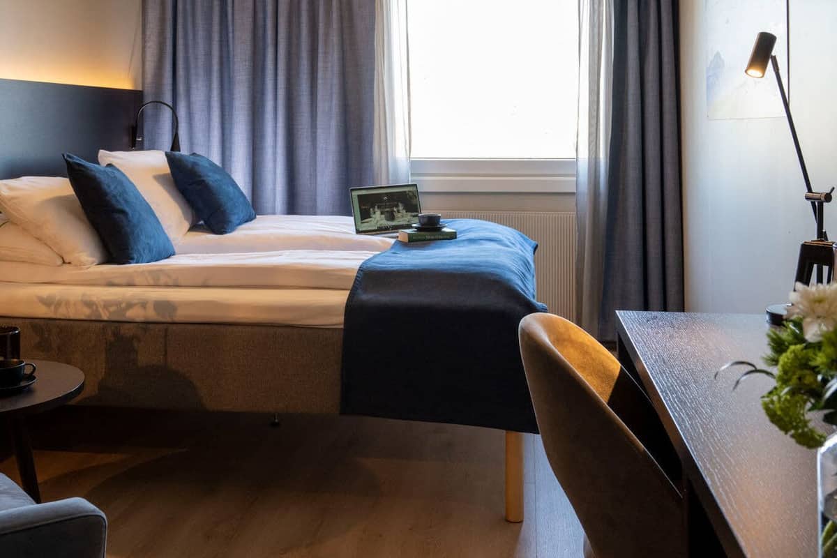 Moderni hotellihuone, jossa on sänky, pieni pöytä, jolla on juoma ja kirja, tuoli sekä työpöytä lampun kanssa.