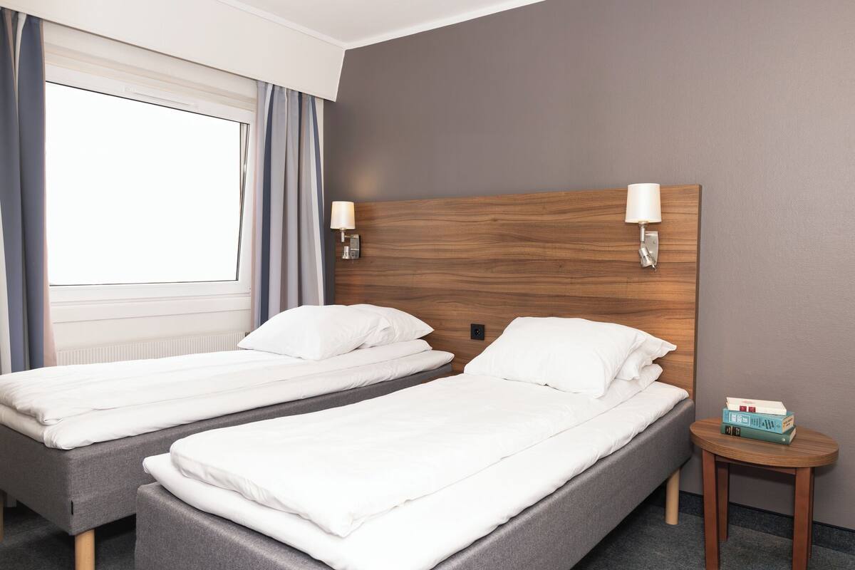 Hotellihuone, jossa on kaksi erillistä sänkyä, pieni puinen yöpöytä ja ikkuna, jossa on verhot.