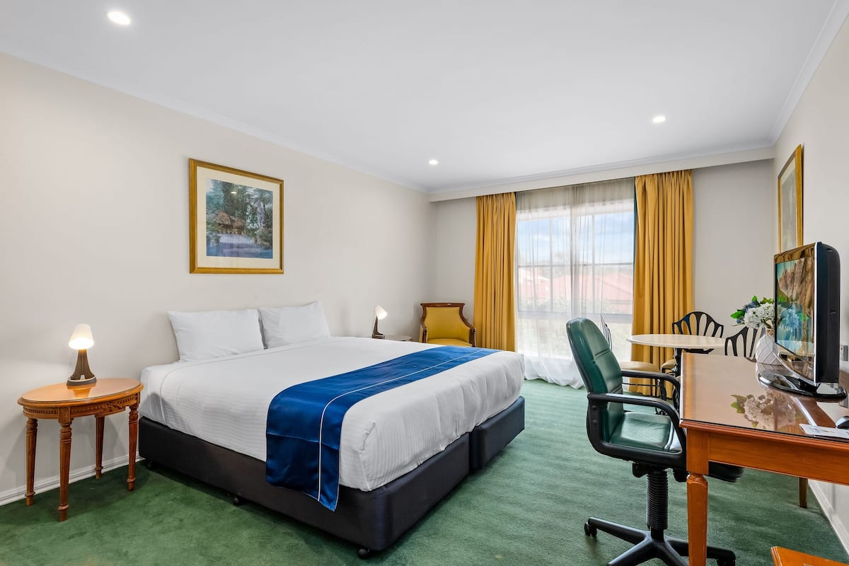 Habitación de hotel con cama, escritorio, silla, lámpara, un cuadro y ventana con cortinas.