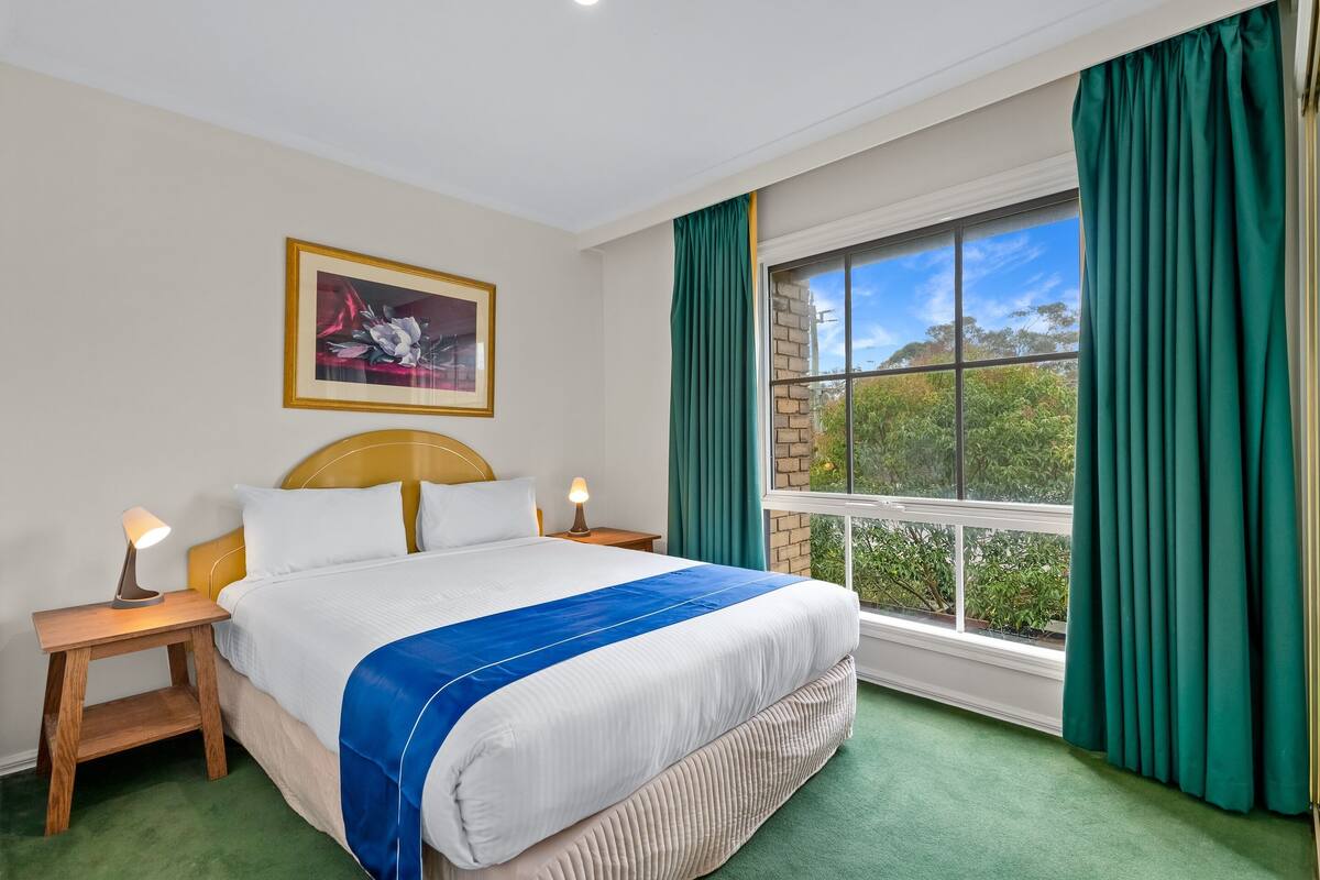 Un cuarto de hotel ordenado con una cama grande, una obra de arte enmarcada en la pared, una ventana con cortinas y vistas a zonas verdes.