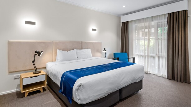 A hotel room with a large bed, a nightstand, a desk, and a window with curtains. - Rowville International Hotel (Rowville)