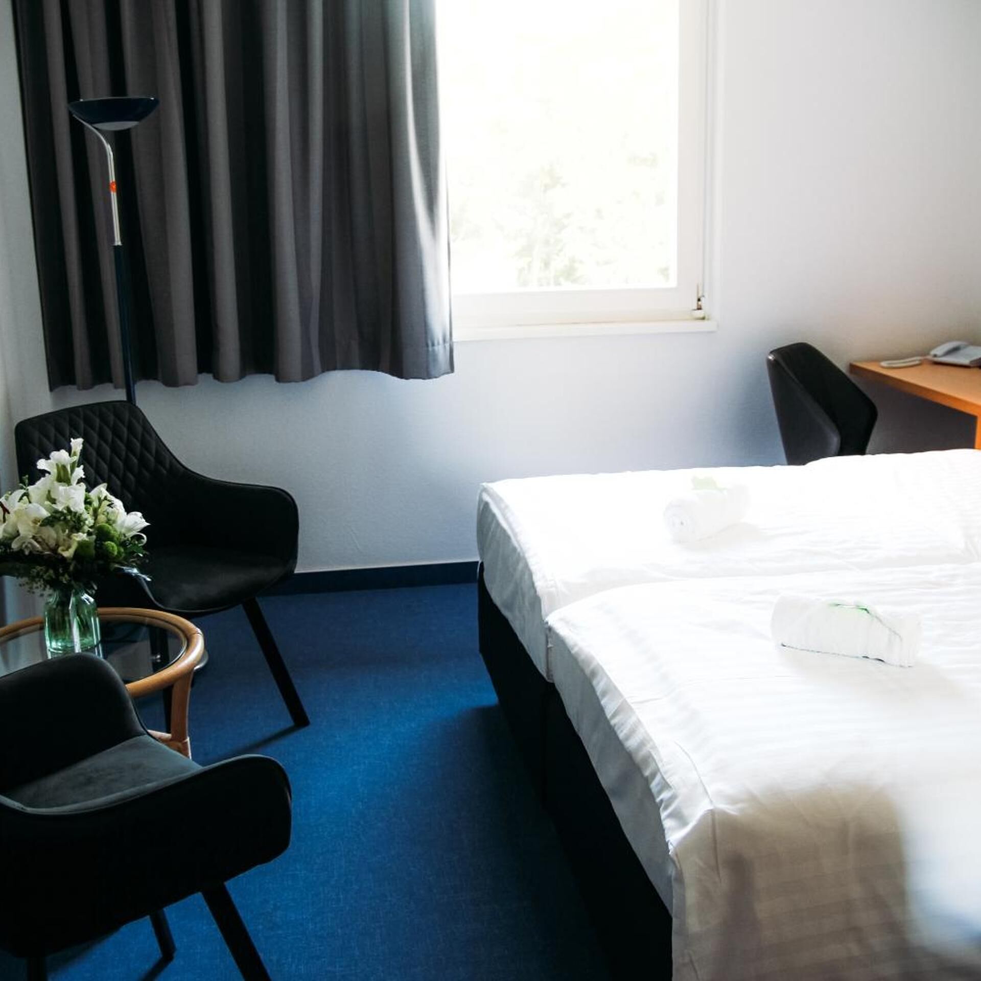 A hotel room with a bed, a desk, a chair, a vase with flowers, and a window with curtains.