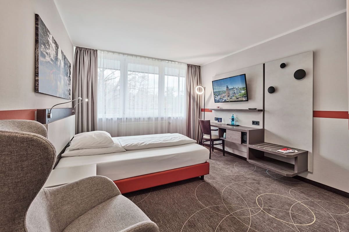 Ein Hotelzimmer mit Bett, Schreibtisch, Stuhl, Fernseher und einem großen Fenster mit Vorhängen.