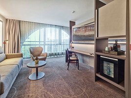 Una habitación de hotel moderna con sofá, dos sillones, una mesita redonda, televisor, cafetera y minibar.