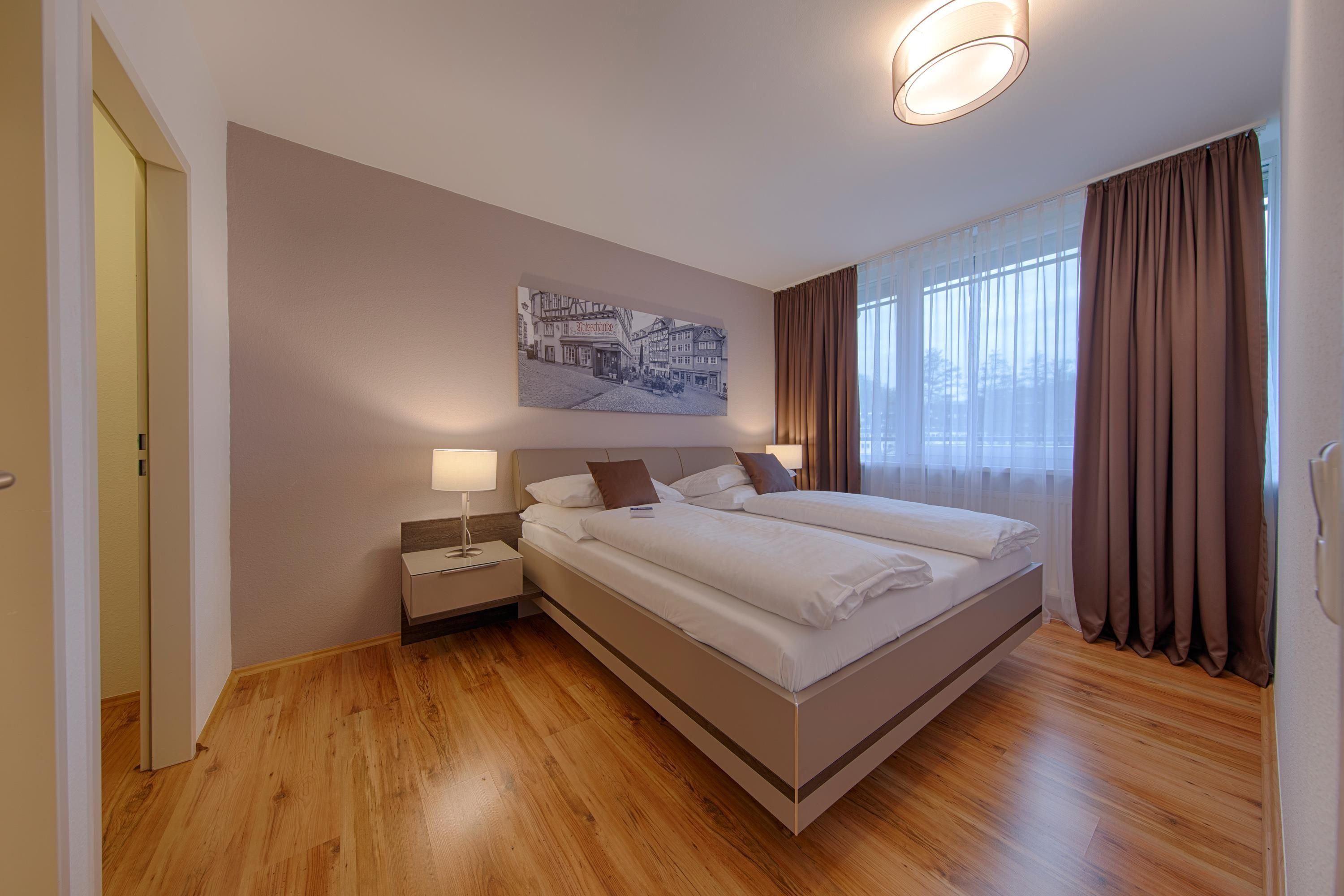 A hotel room with a large bed, a nightstand, a wall-mounted lamp, and a painting of a street scene.