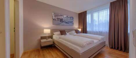 A hotel room with a large bed, a nightstand, a wall-mounted lamp, and a painting of a street scene.