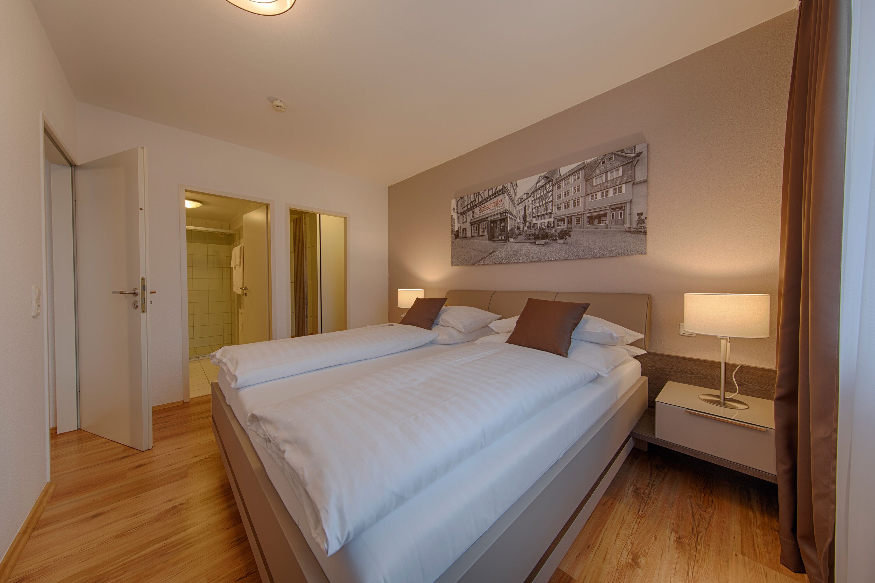 Habitación de hotel con cama doble, mesita de noche con lámpara, un cuadro en la pared y piso de madera.