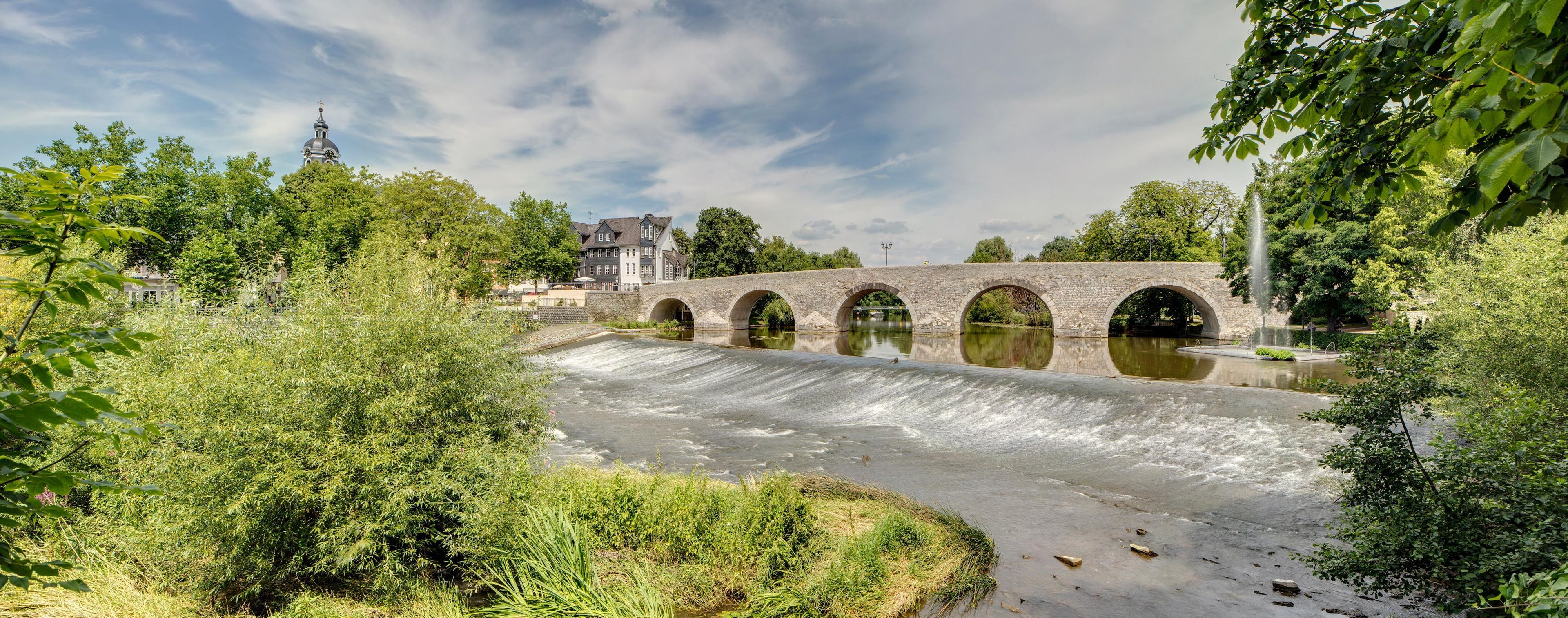 A stone bridge with multiple arches over a river, surrounded by greenery and a building with a tower in the background.