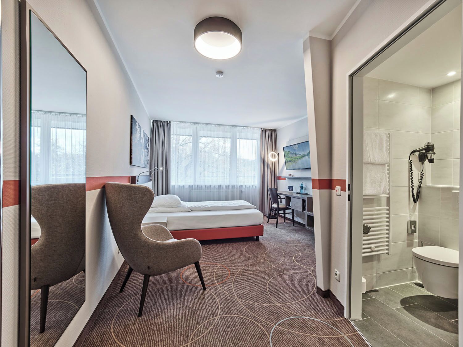 A hotel room with a bed, a desk with a chair, a TV, and a shower area.