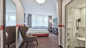 A hotel room with a bed, a desk with a chair, a TV, and a shower area.