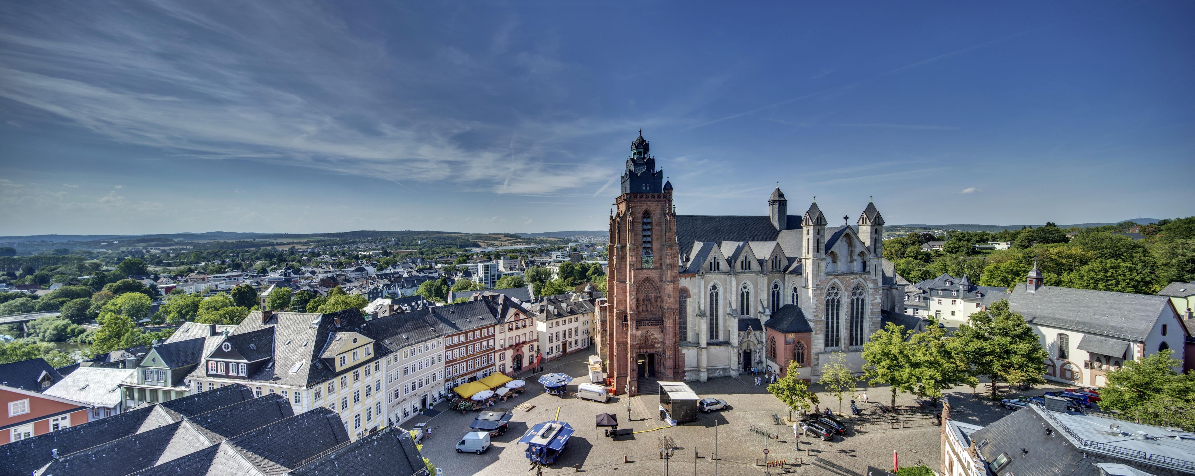Una iglesia histórica con una torre destacada y edificios adyacentes, rodeada por una plaza con coches aparcados y árboles.