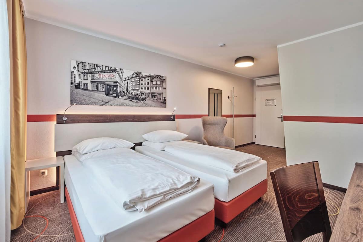 Ein Hotelzimmer mit zwei Betten, einem Schreibtisch, einem Stuhl, einem gerahmten Bild an der Wand und einer Tür.
