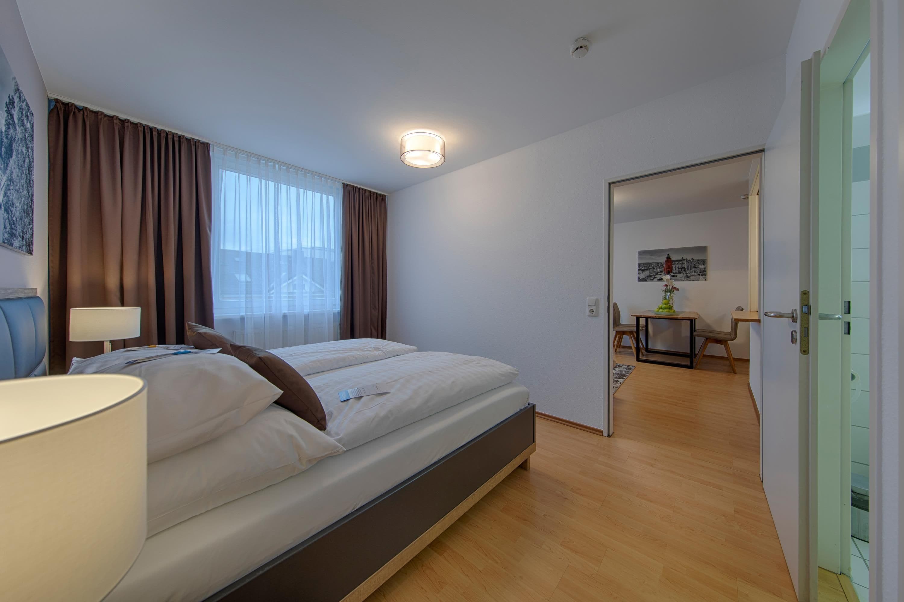 A hotel room with a bed, a desk, a chair, a lamp, and a view of the city through the window.