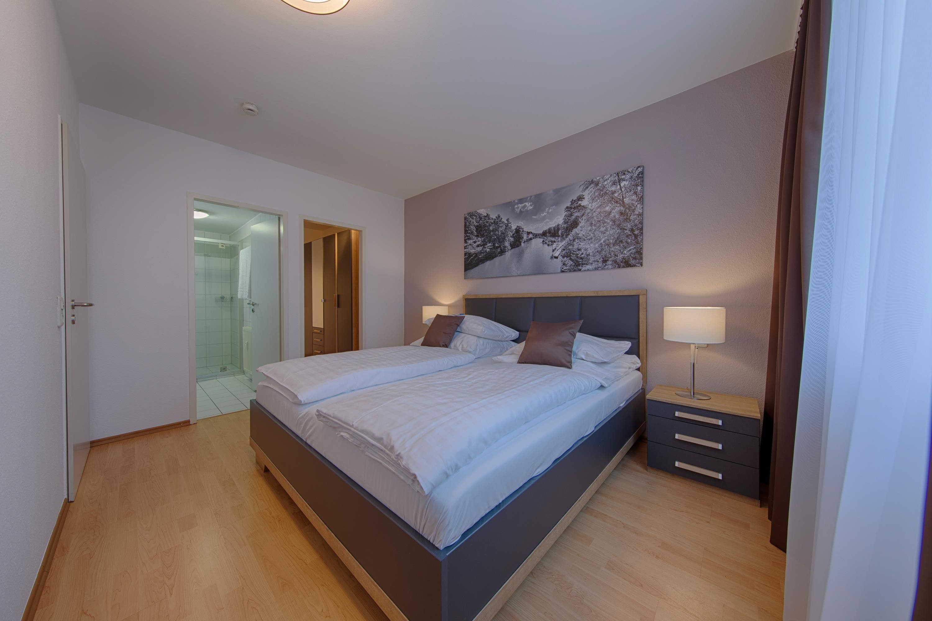 A modern bedroom with a large bed, a bedside table with a lamp, a glass shower enclosure, and a painting on the wall.