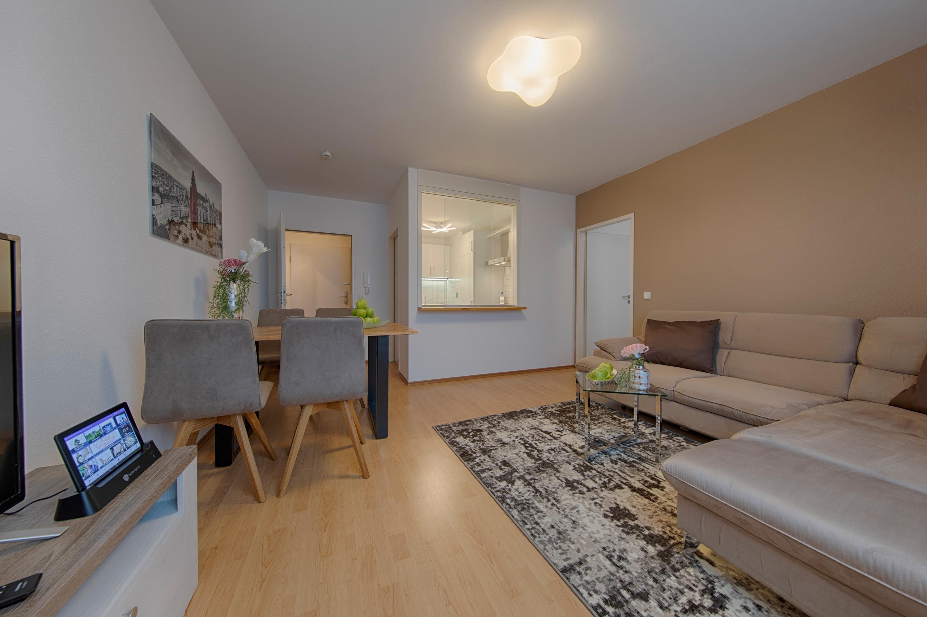 Una sala de estar moderna con un sofá, una zona de comedor con sillas, un televisor y una cocina visible a través de una puerta abierta.