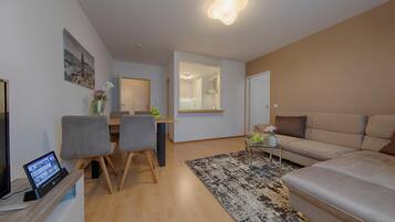 A modern living room with a sofa, a dining area with chairs, a TV, and a kitchen visible through an open door.