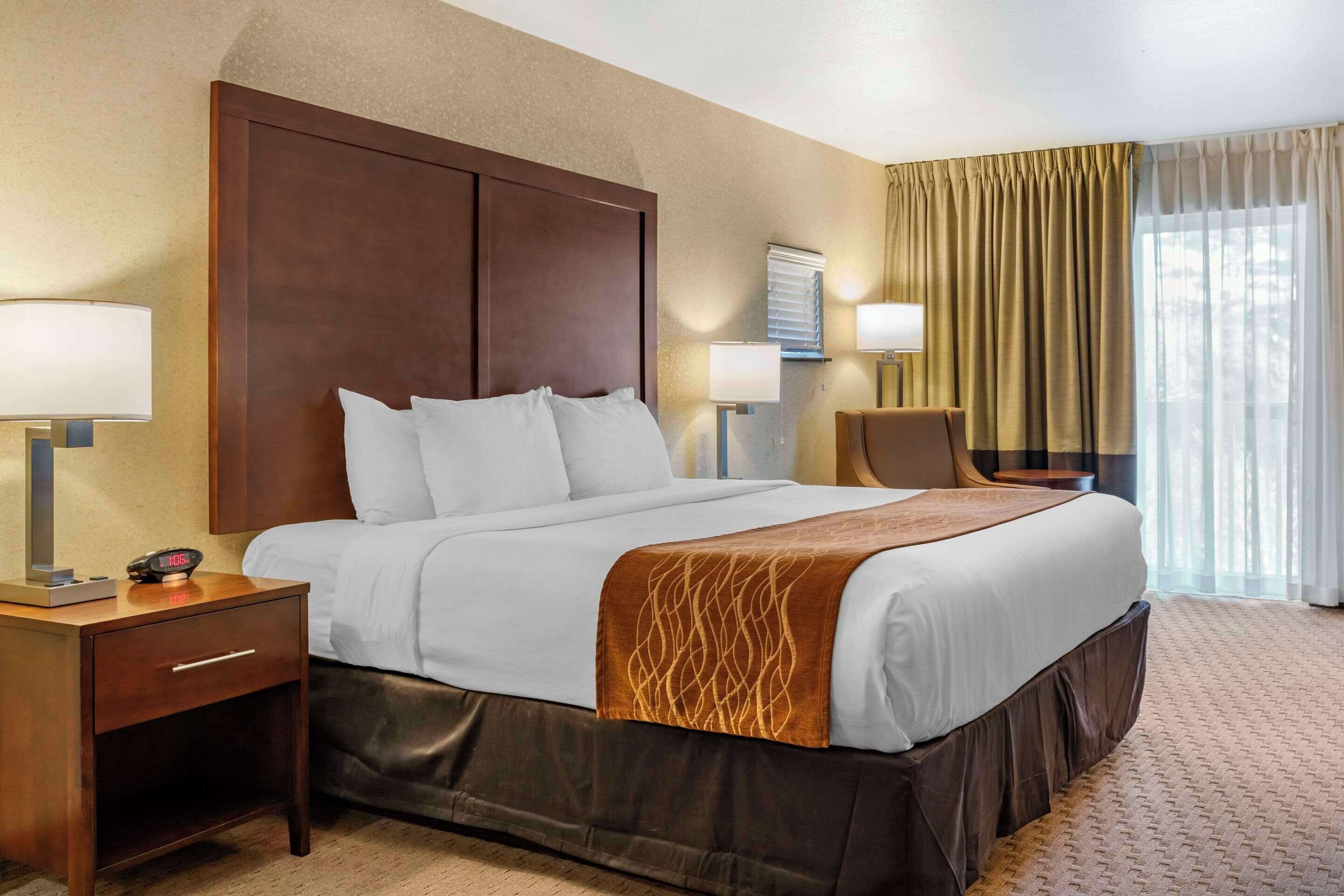 Habitación de hotel con una cama grande, una mesita de noche con lámpara, una silla y un ventanal con cortinas.